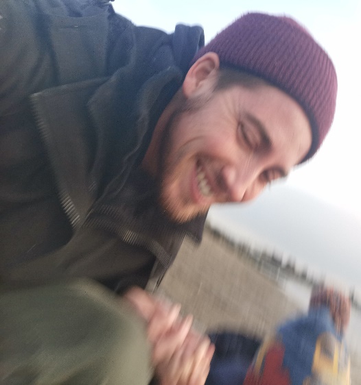 A slightly blurry photo of Gavin smiling widely at the beach looking towards their child, who is out of frame.