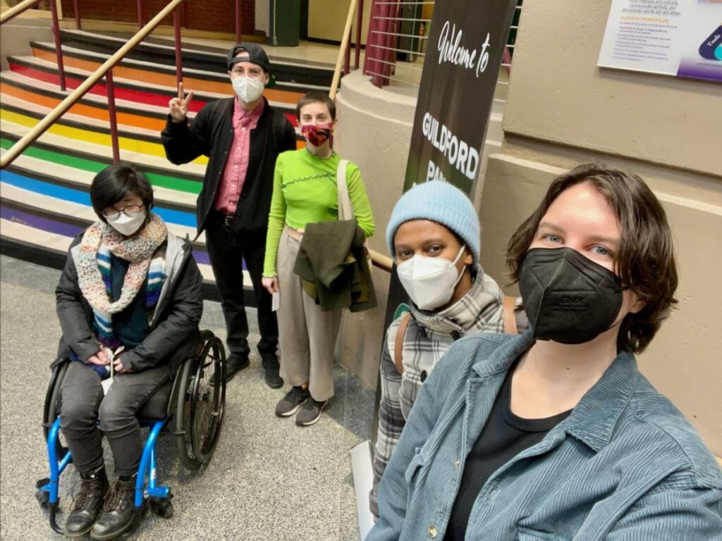 Five people in masks pose for a selfie in front of a rainbow staircase at a school.