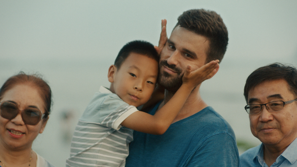 A young Chinese boy squeezes the cheeks of a white man with a beard who is holding him up and posing for a photo with an older Chinese couple.