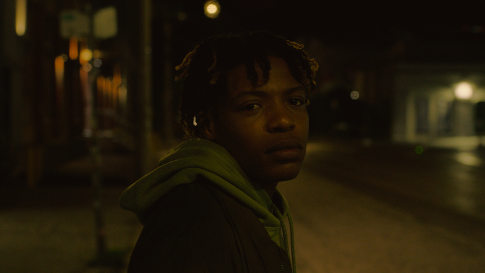 Close up of a black person with short dreads in a hoodie looking behind them on a dark street.