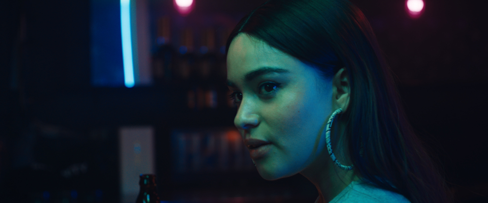 A person with long dark hair and large hoop earrings stands in a bar. There is blue light on their face.